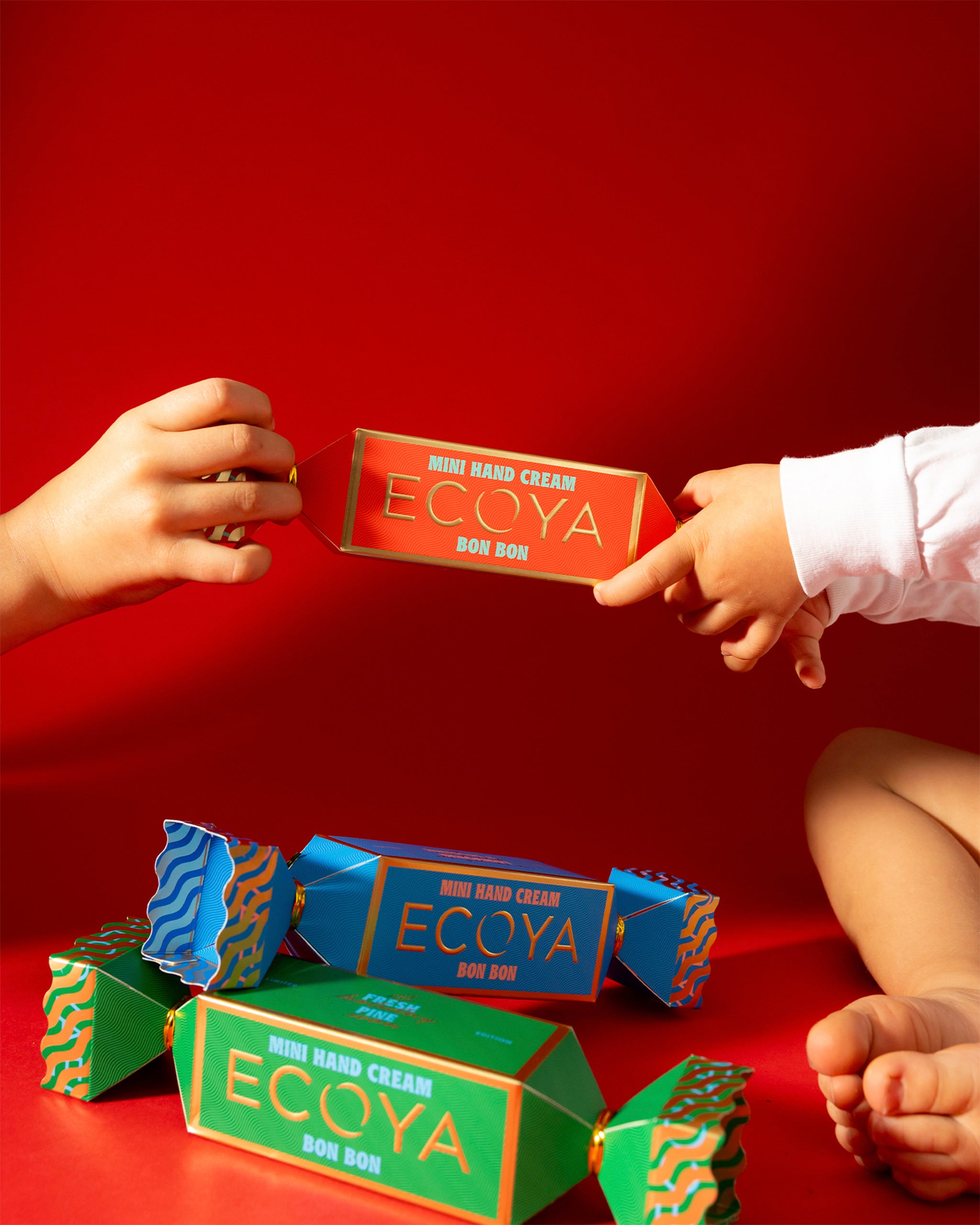 Two hands cracking opening a bon bon of Ecoya hand cream against a red background