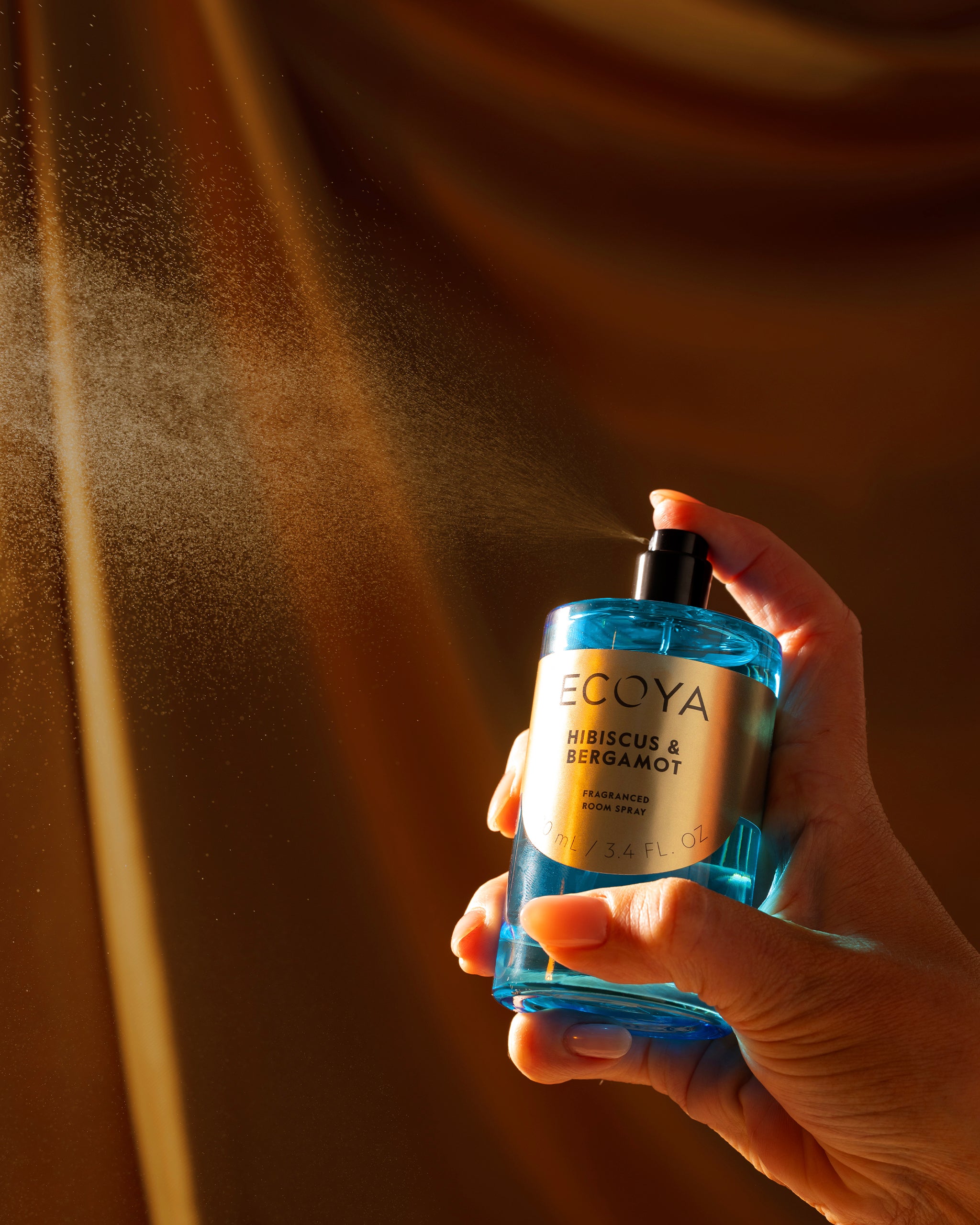 Hand holding a ecoya room spray housed in a blue bottle labeled 'Ecoya' against a brown background