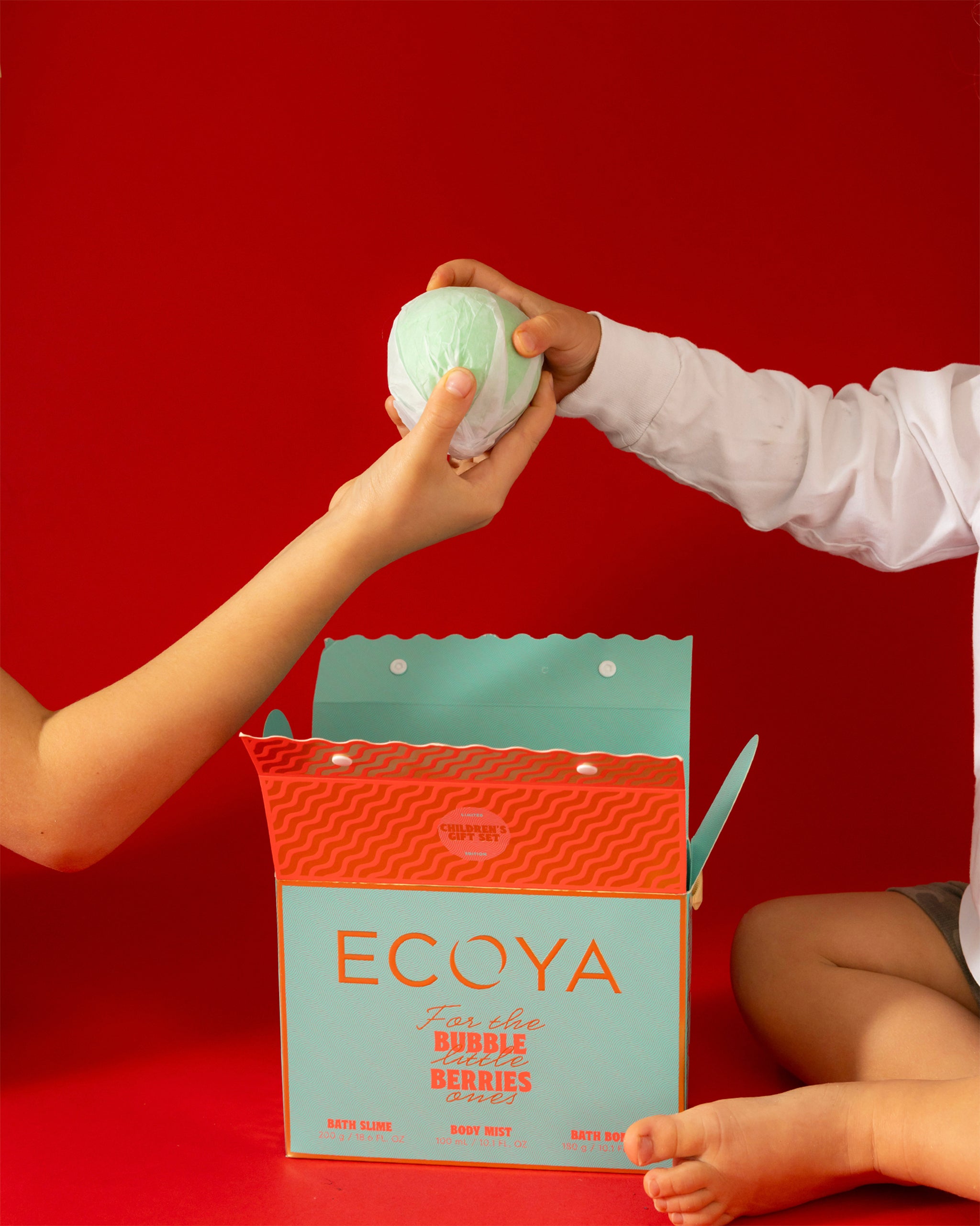 Person exchanging a green bath bomb for an open Ecoya box against a red background