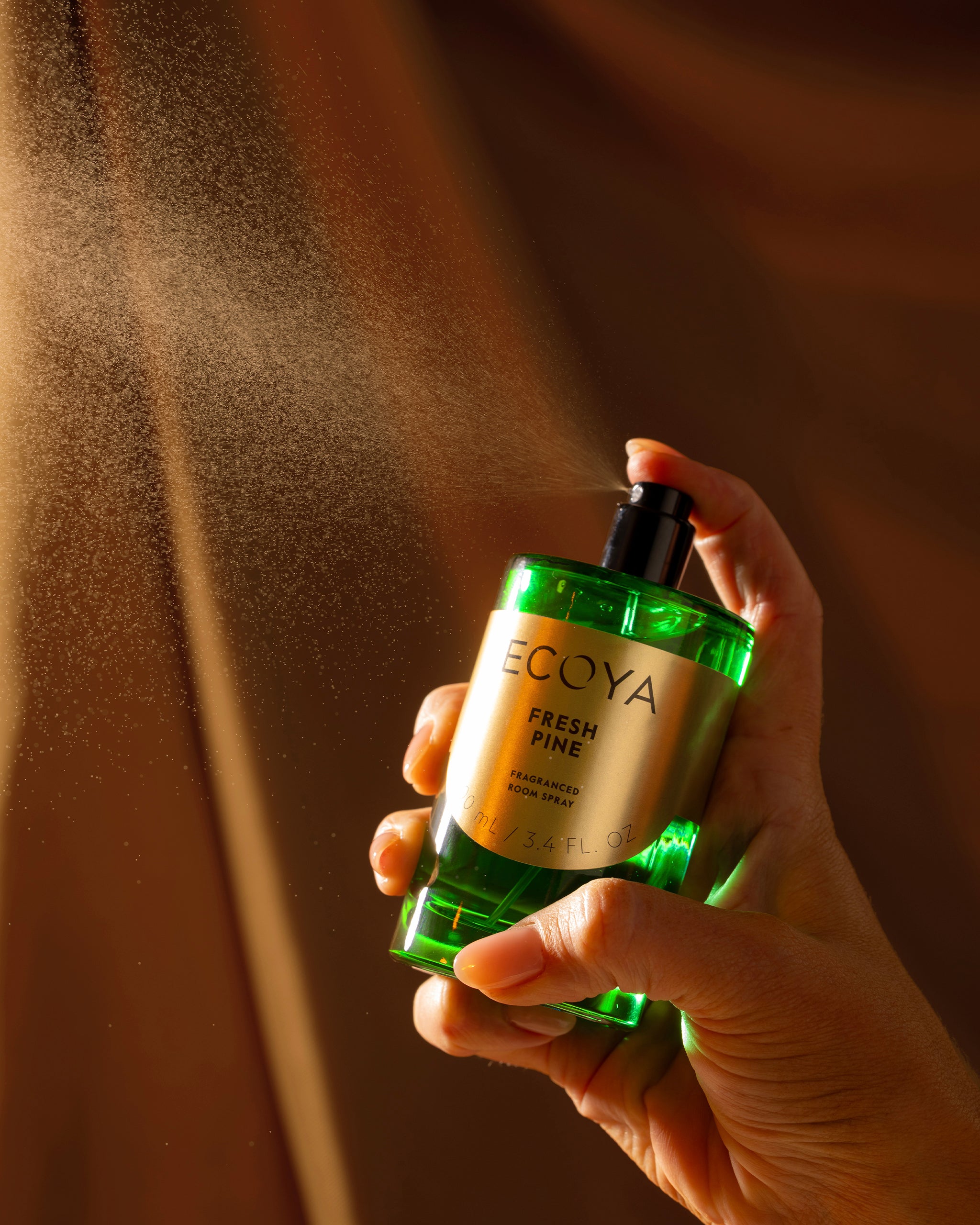 Hand holding a green bottle of Ecoya Fresh Pine room spray with mist being sprayed against a brown background