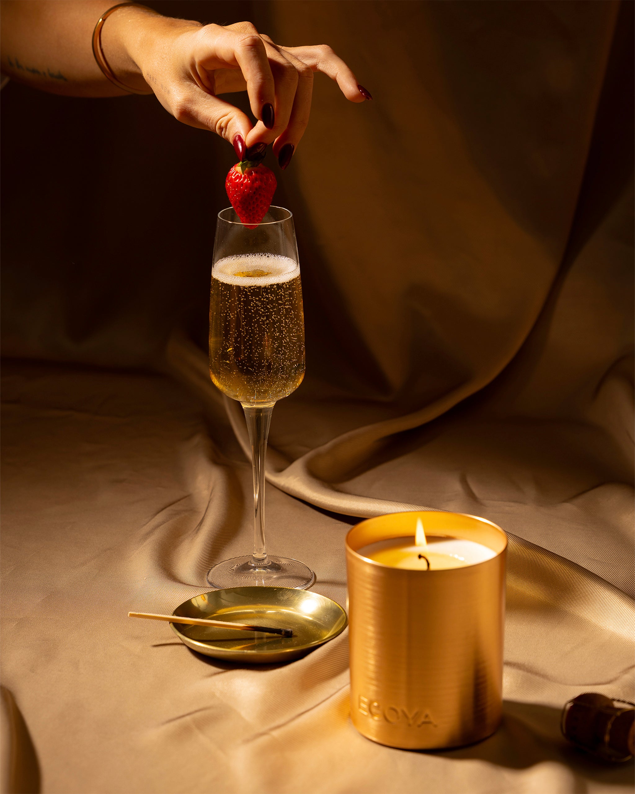 Hand placing a strawberry into a glass of champagne with a lit candle and small dish on a draped fabric background