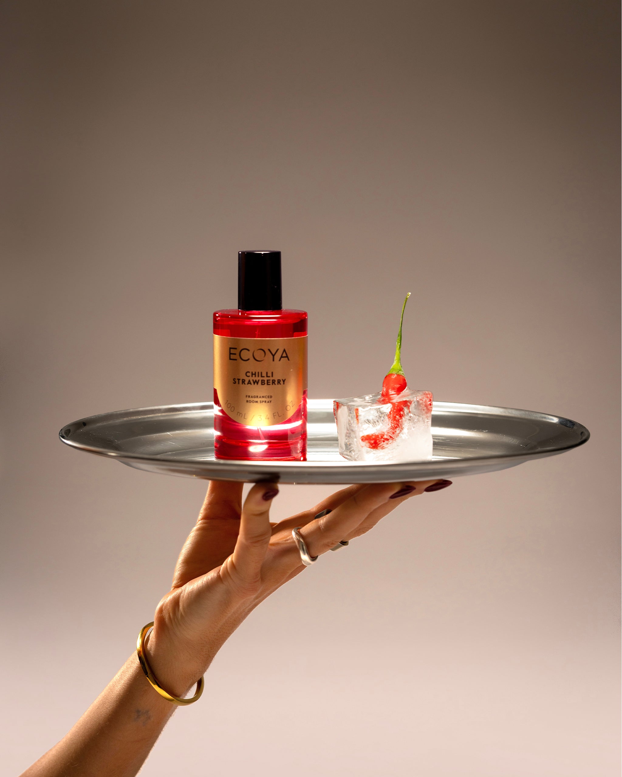 Hand holding a metal tray with a bottle of Ecoya room spray and a small dessert.